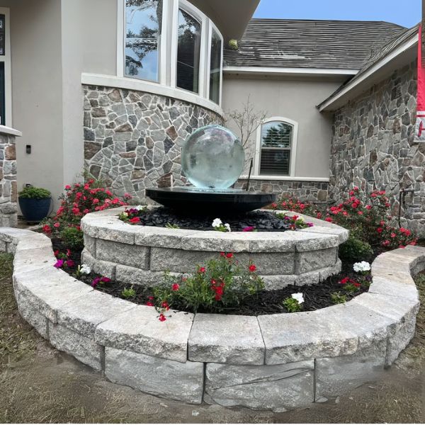 Crystal Sphere Fountain Kit Set in circular stone bed surrounded by flowers in front of house with stone exterior