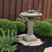 Juliet cast stone birdbath with two decorative birds perched on bowl surrounded by lush green shrubs in a garden setting.